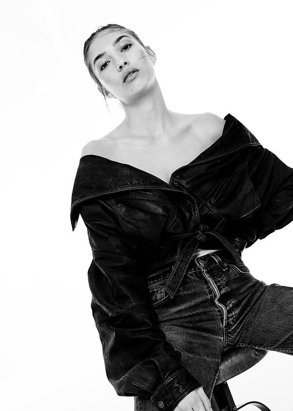 Black and white studio portrait of a young woman in a leather jacket and jeans. Bright, high-key lighting and a neutral background.