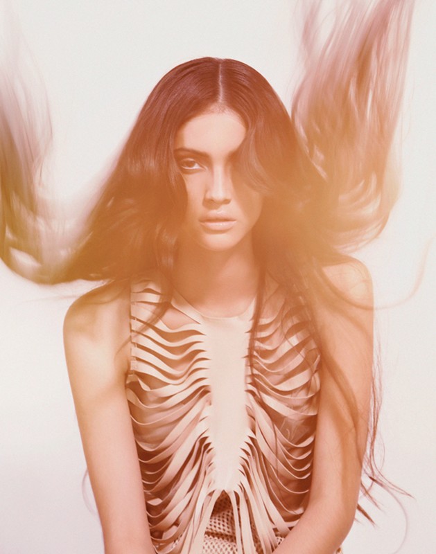 Portrait of a woman with long wavy hair, wearing a beige sleeveless top with a flowing layered design, against a white backdrop.