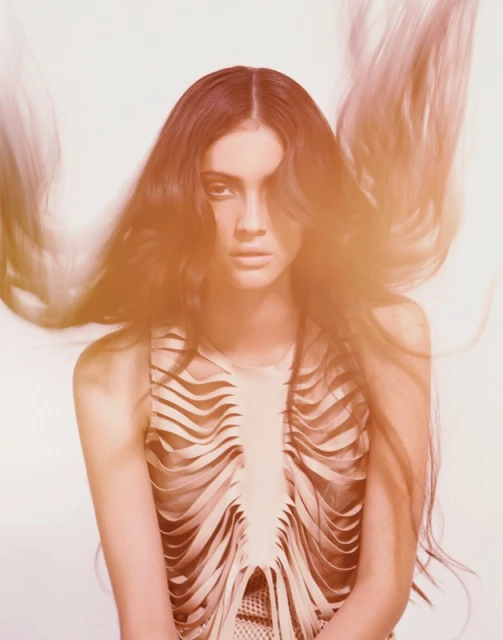 Portrait of a woman with long wavy hair, wearing a beige sleeveless top with a flowing layered design, against a white backdrop.