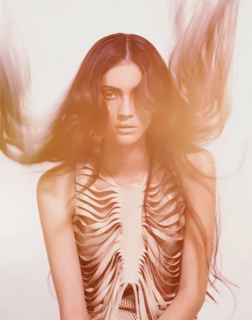 Portrait of a woman with long wavy hair, wearing a beige sleeveless top with a flowing layered design, against a white backdrop.