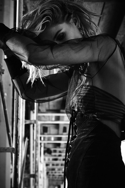 Monochrome portrait of a woman with blonde hair wearing a sheer top, arms crossed, with metal structures in the background.
