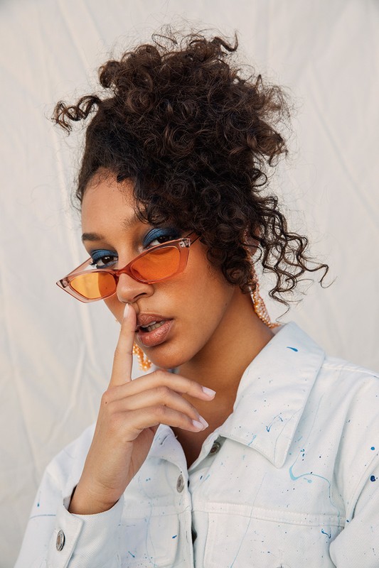 Close-up of a woman with curly hair wearing orange glasses and blue eyeshadow holding a finger to her lips for a portrait.