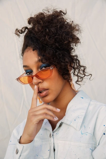 Close-up of a woman with curly hair wearing orange glasses and blue eyeshadow holding a finger to her lips for a portrait.