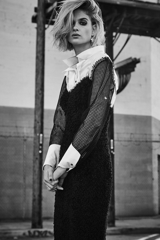 Monochrome portrait of a woman with short blonde hair wearing a black dress and white collared shirt in an urban environment.