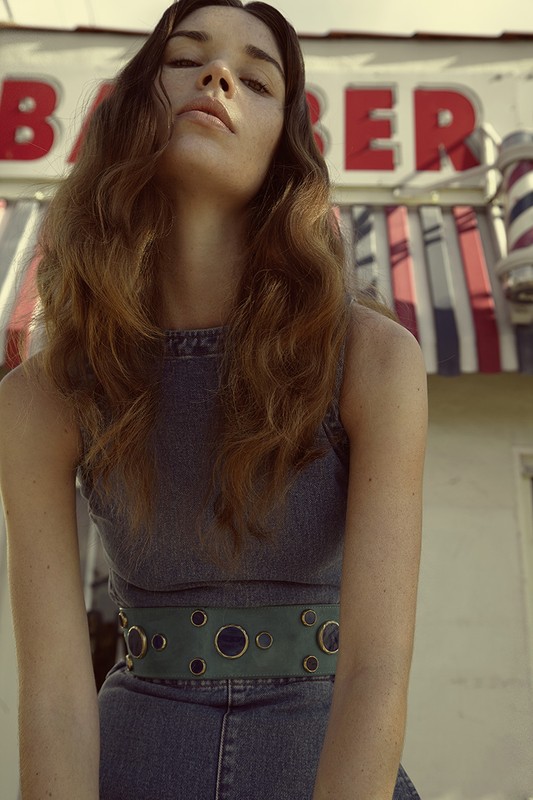 Woman in denim dress and green belt poses in front of barber shop with striped pole, creating a retro, fashionable outdoor shot.