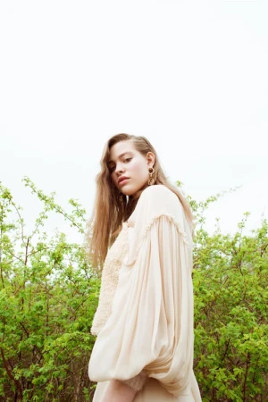 A young woman with long wavy brown hair wearing a cream-colored dress, posed outdoors against a green leafy background.