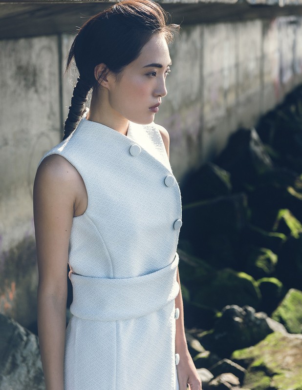 Woman in a sleeveless white dress poses outdoors. A concrete wall and rocks with green moss are visible in the background.