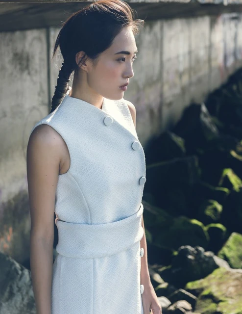 Woman in a sleeveless white dress poses outdoors. A concrete wall and rocks with green moss are visible in the background.