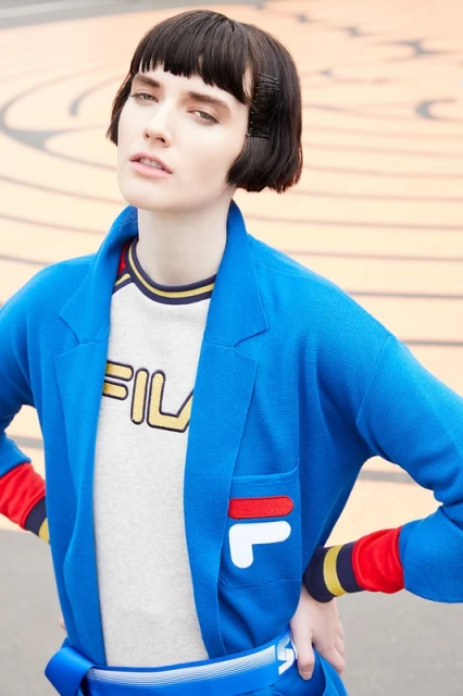 Woman in blue cardigan and white FILA t-shirt poses outdoors against neutral geometric background in a fashion lifestyle image.