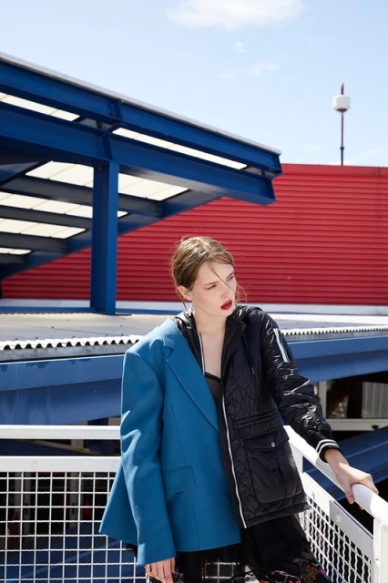 Fashionable woman in a mismatched jacket posing outside with a vibrant blue and red building in the background, blue sky overhead.