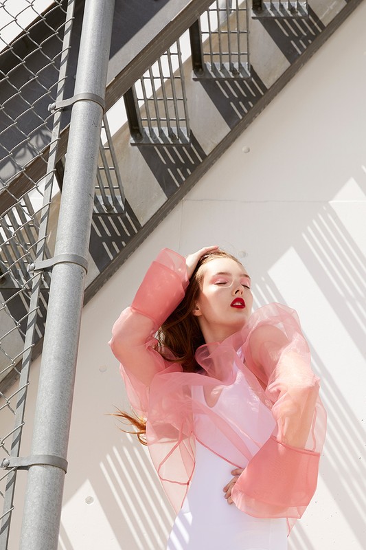 Young woman with red lipstick poses in a sheer pink top with a white tank, next to a metal fence and stairs in sunlight.
