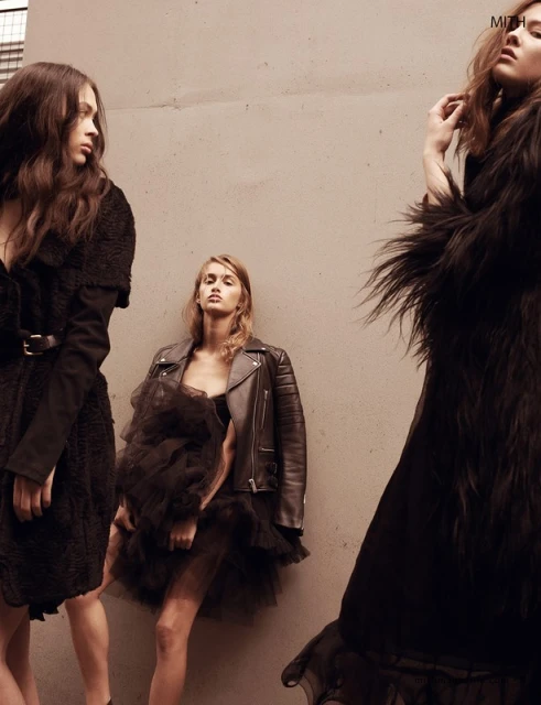 Three female fashion models in black dresses pose against a concrete wall; low angle view in an outdoor setting; neutral color palette.