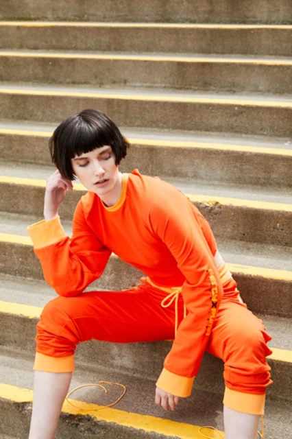 An adult female sits on concrete steps in bright orange apparel. She has dark hair and her head is tilted with her hand near her face.