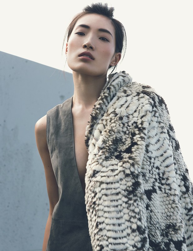 A woman in a grey vest and patterned coat poses against a pale background, with the coat draped over her shoulder.