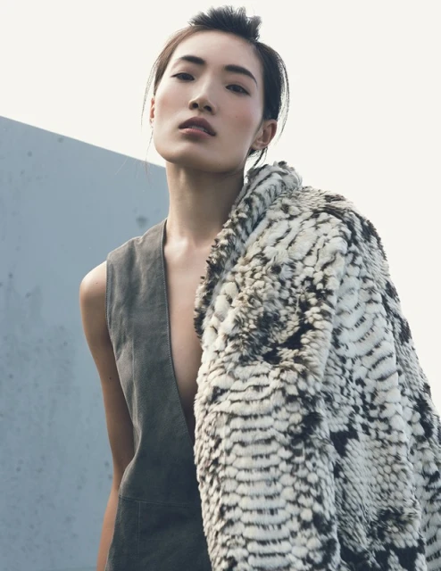 A woman in a grey vest and patterned coat poses against a pale background, with the coat draped over her shoulder.