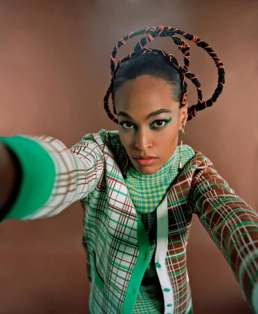 Model with braided sculptural hair and green eyeliner wearing a green and brown plaid cardigan against a warm brown background