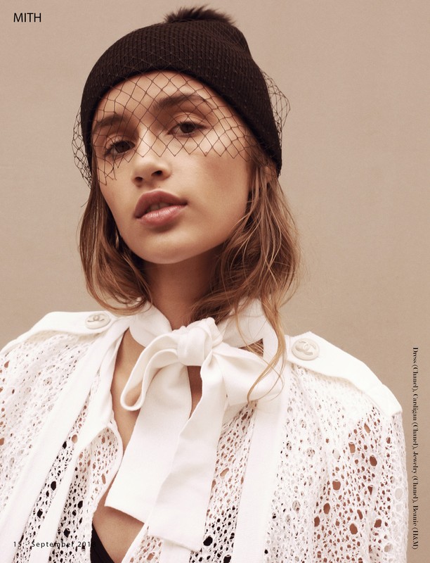 Close-up of a woman with a neutral expression wearing a black knit hat and a white eyelet top against a beige backdrop.