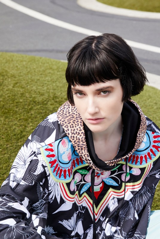 Young woman with short dark hair poses in an outdoor setting wearing a patterned hoodie with a leopard-print hood.