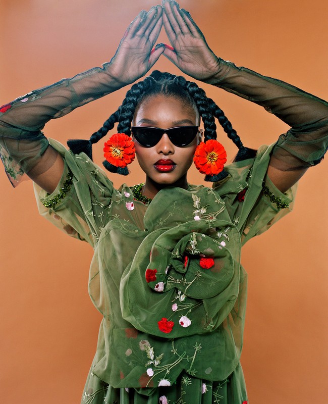 Glamorous young woman in sunglasses with braids, decorative orange hair accents, sheer sleeves, and floral patterned blouse against solid background.