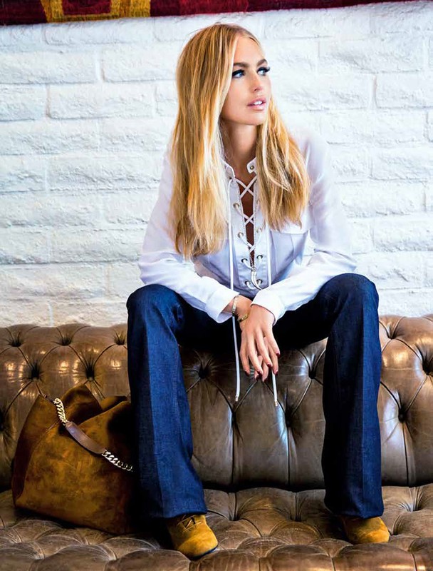 Blonde woman in jeans and white shirt sits on a brown leather couch indoors with a brown tote bag on the couch next to her.