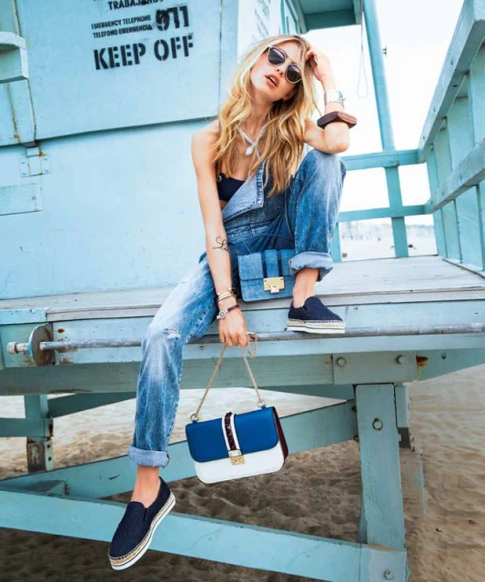 A blonde woman in jeans and sunglasses sits on a lifeguard tower at the beach holding a purse. The sky is light blue.