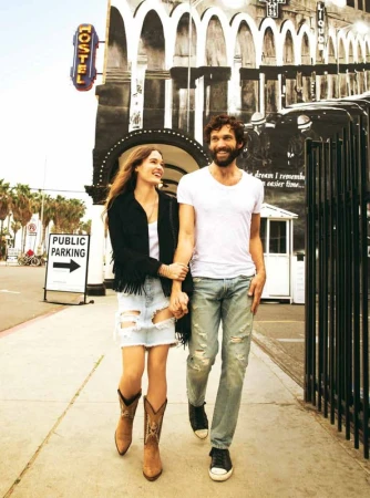 A smiling couple walks hand-in-hand on a sidewalk in front of a building with an old mural, enjoying a sunny day out together.