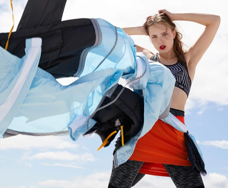Fashion editorial shot of a woman outdoors in sportswear with her hands in her hair, and apparel in motion against a blue sky.