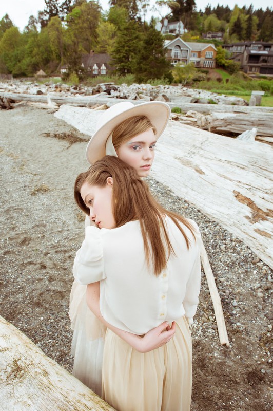 Two young women embrace on a sandy beach with driftwood. The woman in front rests her head on the other's back, wearing cream-colored clothing.