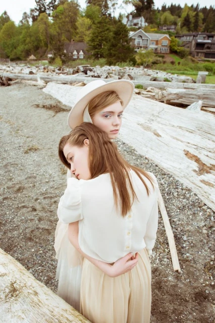 Two young women embrace on a sandy beach with driftwood. The woman in front rests her head on the other's back, wearing cream-colored clothing.