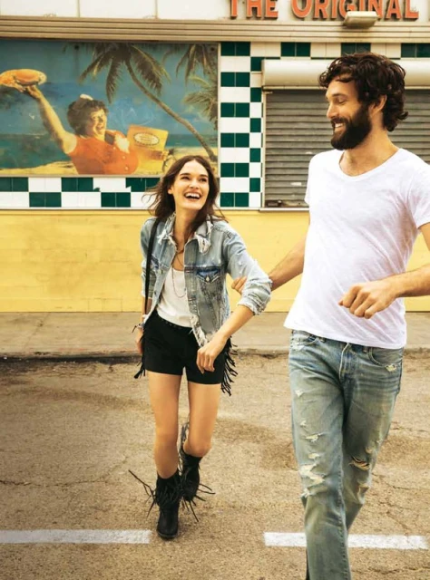 A laughing couple walks on a road outside a diner with a vintage mural. The woman wears a denim jacket and fringe shorts. The man is in jeans and a white t-shirt.