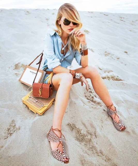 Blonde fashion model in sunglasses sits on a sand dune wearing a denim shirt and shorts with handbags and gladiator sandals.