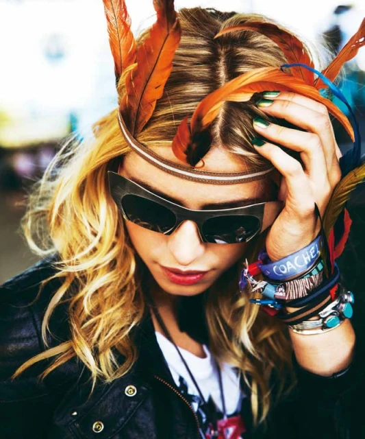 Fashionable young woman wearing sunglasses and a feathered headband poses indoors. She wears a black leather jacket with metal studs.