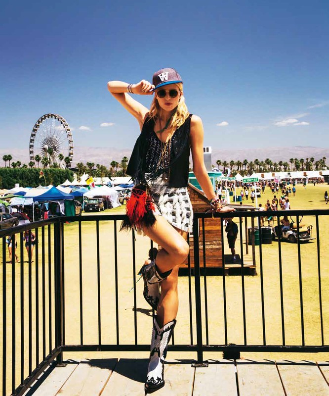 A young woman stands on a platform overlooking a music festival with a Ferris wheel in the background on a sunny day.