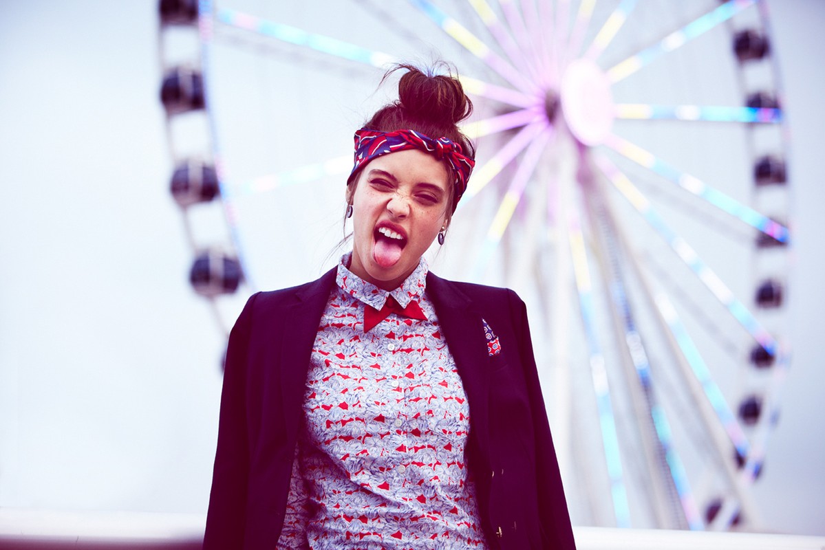 Young woman in blazer playfully sticks her tongue out at the camera against a backdrop featuring a blurred Ferris wheel.