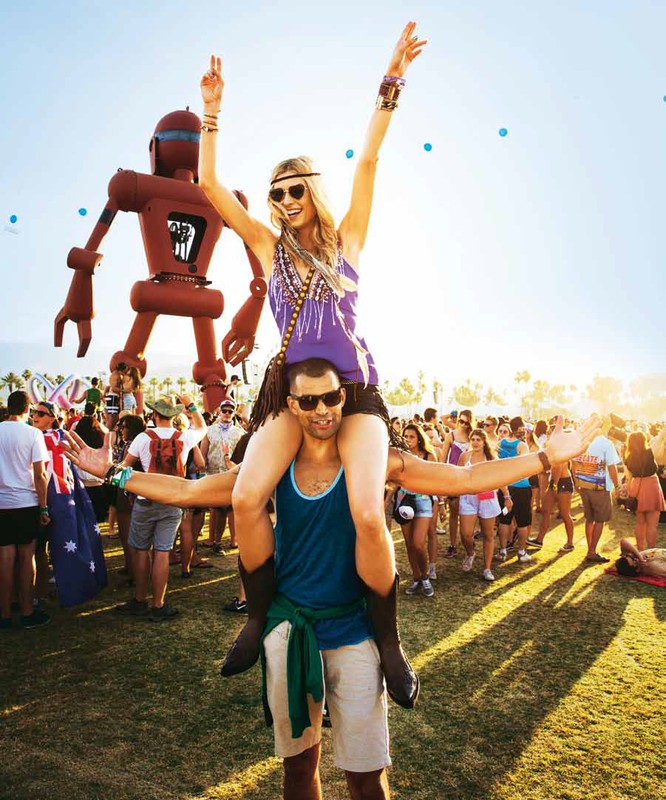 Woman on man's shoulders at an outdoor music festival with a large robot sculpture and crowd in the background, sunny weather.