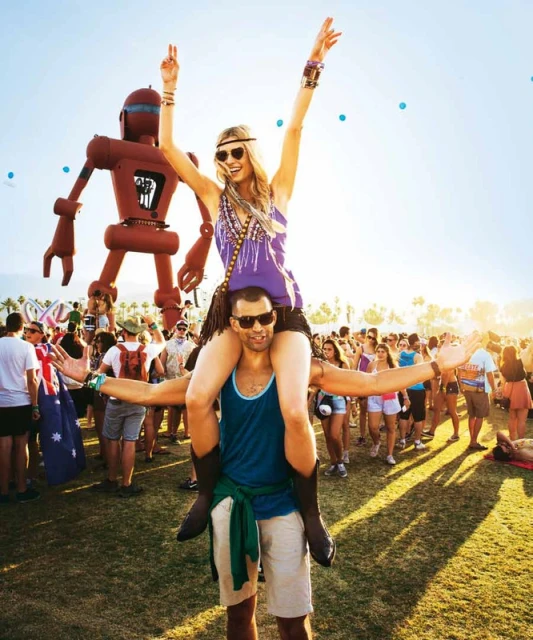 Woman on man's shoulders at an outdoor music festival with a large robot sculpture and crowd in the background, sunny weather.