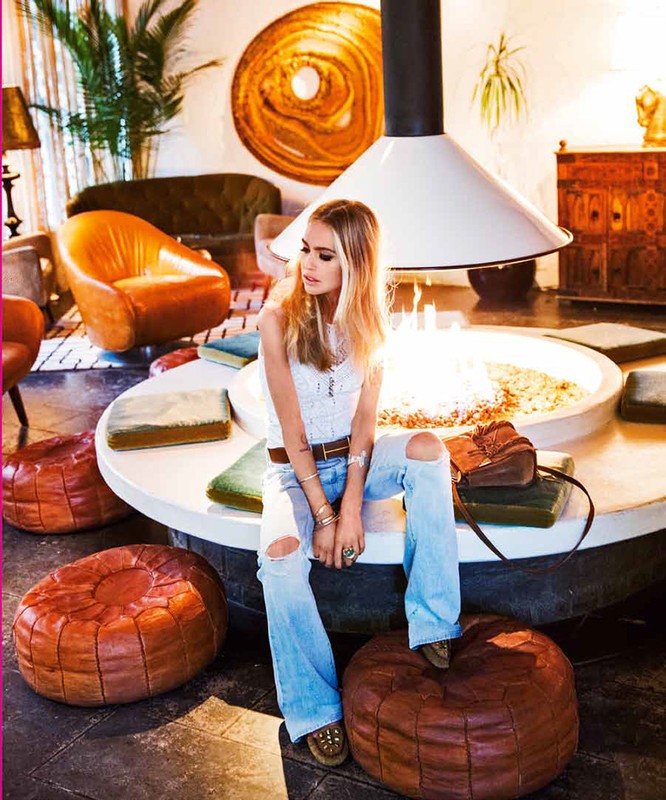 Woman with long blonde hair sitting indoors on a circular structure with a white top and brown accents, wearing light denim jeans.