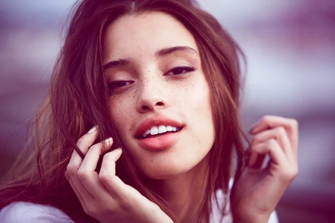 Close-up portrait of a young adult woman with freckles, medium brown hair, fair skin, and pink lips, smiling and holding her hair.