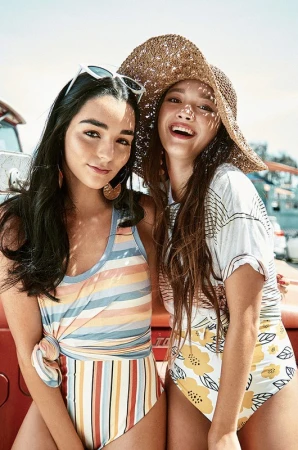 Two smiling young women standing outdoors together, one wearing a straw sun hat and patterned top.