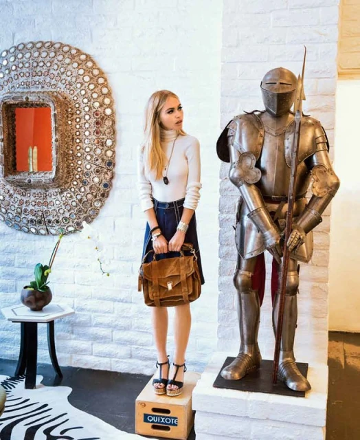 Fashion shot of a blonde adult female posing on a box next to a knight's armor, holding a brown handbag in an indoor setting.