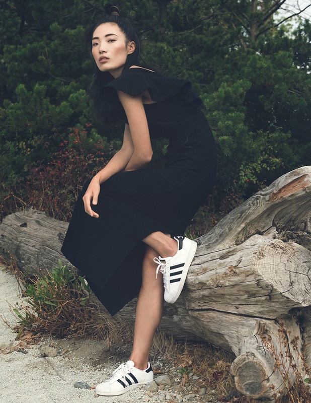 Asian woman in a black dress and sneakers sitting on a log in a forest. Fashion editorial style with natural lighting and outdoor setting.