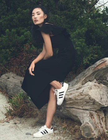 Asian woman in a black dress and sneakers sitting on a log in a forest. Fashion editorial style with natural lighting and outdoor setting.