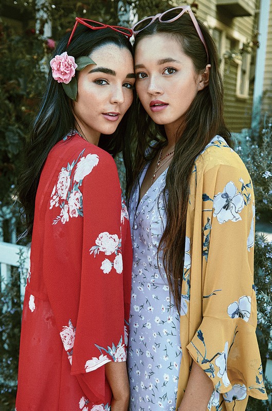 Two young women stand closely together outside. One wears a red floral kimono and glasses, the other a floral dress and yellow kimono.
