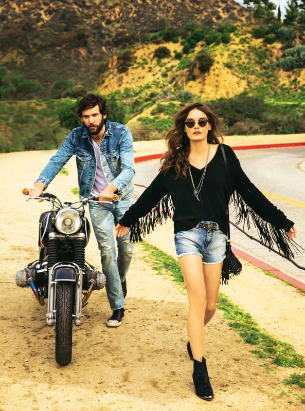 A woman walking in front of a man pushing a motorcycle along a dirt road with a hillside background.