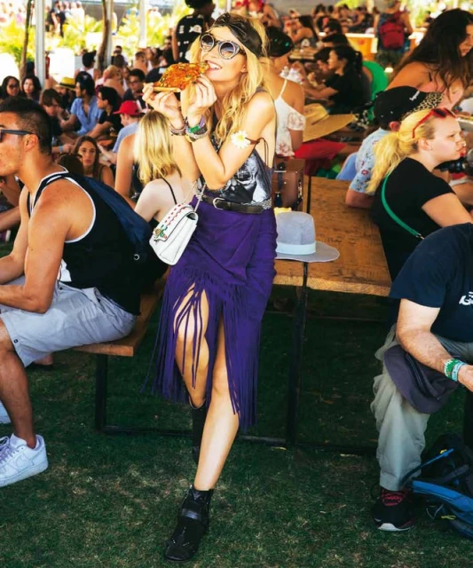 Blonde woman wearing sunglasses and a fringed skirt, holding a slice of pizza at an outdoor festival. Crowd visible in background.
