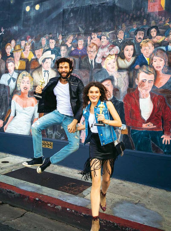 A man and woman holding awards jump with a mural of classic Hollywood stars in the background, capturing a fun, candid moment.