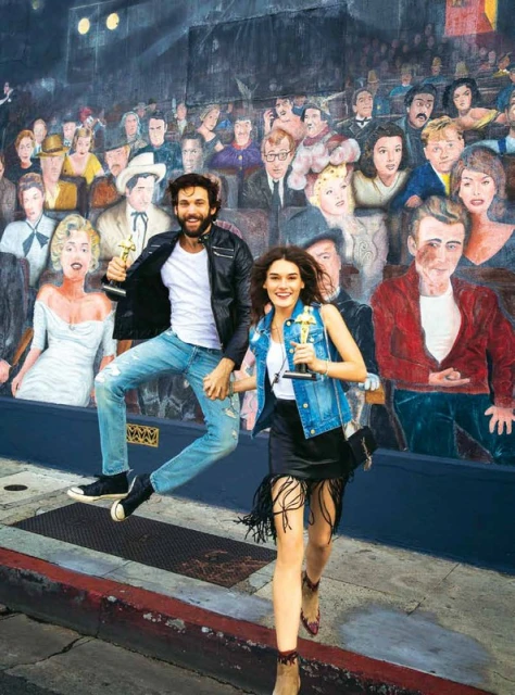 A man and woman holding awards jump with a mural of classic Hollywood stars in the background, capturing a fun, candid moment.