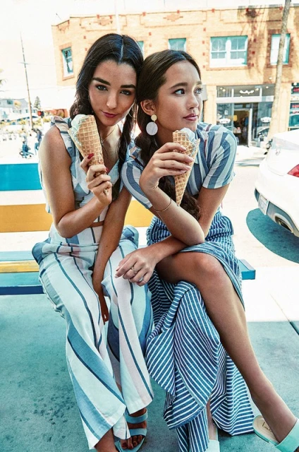 Two young women sit outdoors on a bench wearing striped summer clothing and holding ice cream cones on a sunny day near buildings.