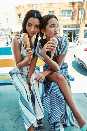 Two young women sit outdoors on a bench wearing striped summer clothing and holding ice cream cones on a sunny day near buildings.