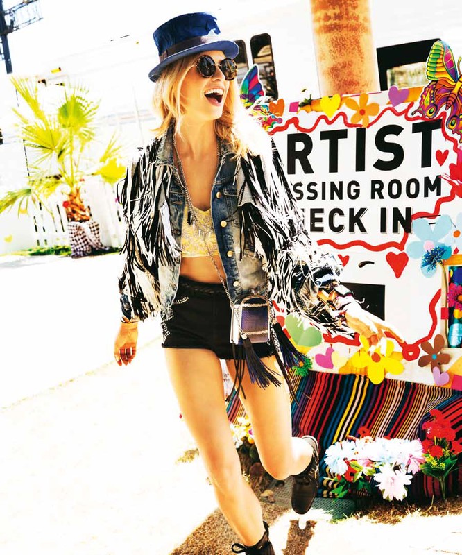 A smiling woman in a fringe jacket, hat, and sunglasses walks past an "Artist Dressing Room" sign on a sunny day outside.